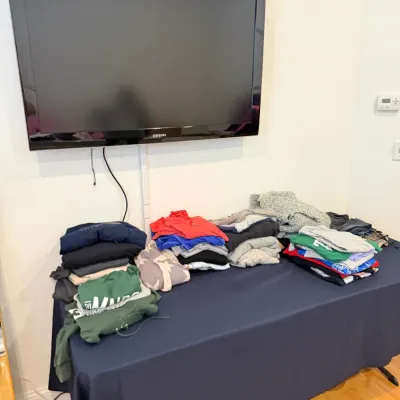 Sale Photo Thumbnail #64: A wall-mounted Samsung flat-screen television above a table arranged with stacks of folded casual clothing and sweatshirts.
