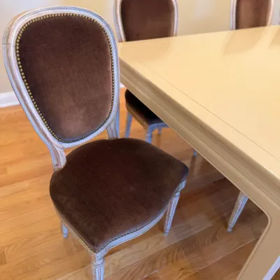 Sale Photo Thumbnail #17: French Provincial style dining chairs featuring whitewashed wooden frames, brown velvet upholstery, and brass nailhead trim surrounding a large dining table.
