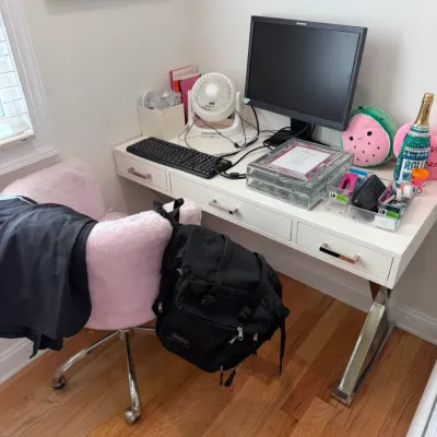 Sale Photo Thumbnail #98: Modern white writing desk with chrome X-legs paired with a pink faux-fur swivel office chair, alongside a Lenovo computer monitor and desk accessories.