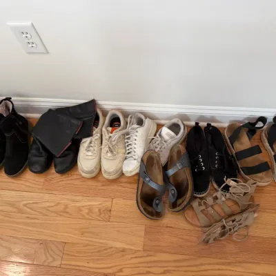Sale Photo Thumbnail #56: Collection of women's shoes and sandals including black ankle boots, sneakers, Birkenstock sandals, espadrilles, and strappy sandals lined up on a hardwood floor.