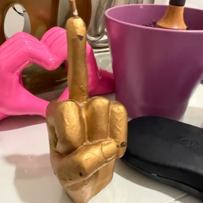 Sale Photo Thumbnail #81: Novelty gold middle finger candle displayed alongside decorative tabletop items, including a pink heart-hands sculpture and a purple planter.