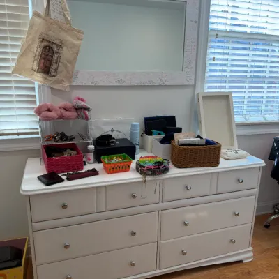 Sale Photo Thumbnail #102: White wooden six-drawer dresser topped with miscellaneous personal items, storage baskets, and accessories, paired with a matching wall mirror above.