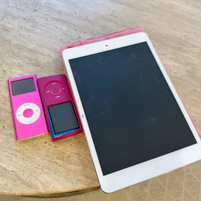 Sale Photo Thumbnail #85: Collection of Apple electronics including a white iPad mini in a pink case alongside two pink iPod Nano devices, circa 2000s to 2010s.