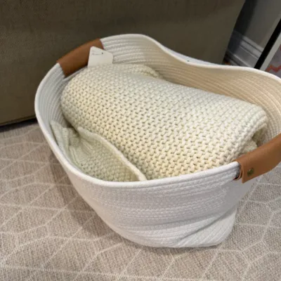 Sale Photo Thumbnail #11: White woven rope storage basket with brown faux leather handles, filled with a folded cream-colored chunky knit blanket.
