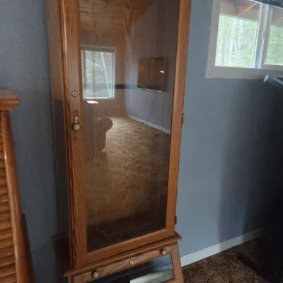 Sale Photo Thumbnail #81: Tall oak display cabinet featuring a full-length glass-front door and bottom drawer storage, showing prominent wood grain and vintage brass hardware.