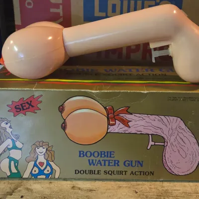 Sale Photo Thumbnail #29: Vintage plastic novelty 'Boobie Water Gun' gag item, shown resting atop its original printed cardboard box with retro illustrated graphics.