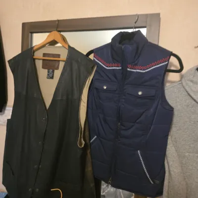 Sale Photo Thumbnail #119: Two men's vests hanging on a mirror, including a black leather snap-front vest and a blue padded zip-up vest with embroidered shoulder trim.