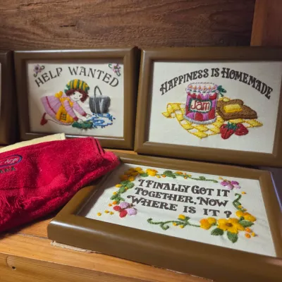 Sale Photo Thumbnail #151: Collection of vintage framed embroidery sampler art featuring cheerful domestic sayings, displayed alongside a folded red souvenir towel.