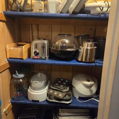 Sale Photo Thumbnail #112: Pantry shelves filled with various small kitchen appliances and cookware, including a stainless steel toaster, West Bend Stir Crazy popcorn popper, blender, food processor, muffin tins, and baking sheets.