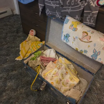 Sale Photo Thumbnail #120: Open vintage blue metal footlocker trunk filled with vintage baby clothes, blankets, and textiles, accompanied by a nearby doll and Pendleton-style jacket.