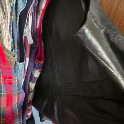 Sale Photo Thumbnail #146: Black leather jacket hanging among various vintage garments, including a plaid shirt and knit vest, inside a closet.