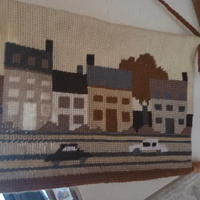 Sale Photo Thumbnail #153: Mid-century vintage latch hook wall hanging depicting a street scene with buildings and cars, mounted on wooden dowels with a hanging cord.