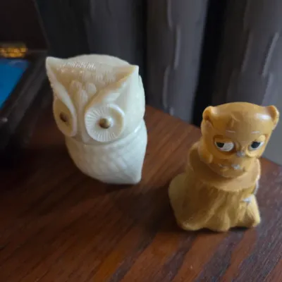 Sale Photo Thumbnail #106: Pair of vintage owl figurines, including one carved off-white plastic or resin owl with brass-toned eye accents and a small yellow composition owl perched on a tree stump.