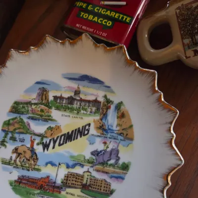 Sale Photo Thumbnail #105: Vintage Wyoming souvenir plate with gold scalloped rim, displayed alongside a red Velvet tobacco pocket tin and a ceramic mug.