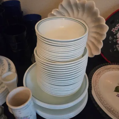 Sale Photo Thumbnail #113: Large stack of white Corelle dishware with green banded trim, displayed alongside various ceramic plates and dark glassware.