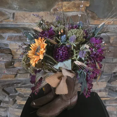 Sale Photo Thumbnail #80: Rustic floral arrangement set in brown leather cowboy boots, featuring sunflowers, purple blossoms, dried greenery, and a burlap bow accent.