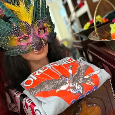Sale Photo Thumbnail #34: Feathered masquerade mask with sequin trim displayed on a mannequin head, alongside a folded Foreigner graphic band t-shirt.