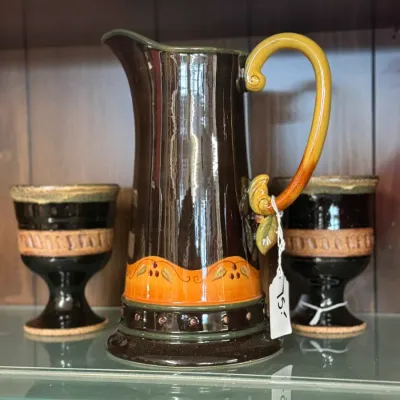 Sale Photo Thumbnail #32: Glazed ceramic pitcher with yellow-orange accent band and handle, displayed alongside two matching goblets on a glass shelf.