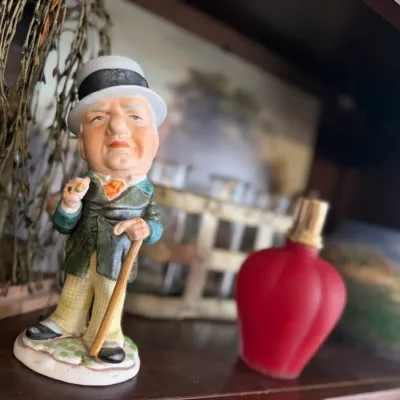 Sale Photo Thumbnail #7: Vintage painted ceramic figurine of W.C. Fields holding a cane and cigar, displayed alongside a red glass perfume bottle on a wooden shelf.