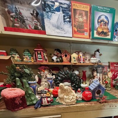 Sale Photo Thumbnail #2: Collection of festive holiday decor and collectibles including boxed Pfaltzgraff items, ceramic nativity scenes, snowmen figurines, and Christmas tree decorations displayed on wooden shelves.