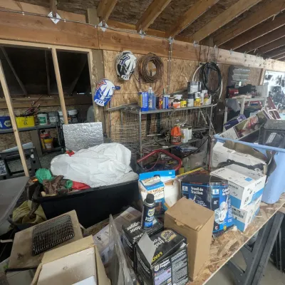 Sale Photo Thumbnail #10: Workshop workbench filled with assorted household and garage items, including boxed plumbing fixtures, insect fogger packs, bird feeders, and Fox motocross helmets hanging on the wall.