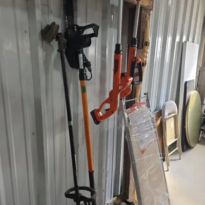 Sale Photo Thumbnail #114: Collection of lawn power tools hanging on a wall, including string trimmers and a pole saw, alongside folding chairs.