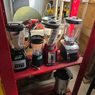 Sale Photo Thumbnail #120: Collection of assorted electric kitchen blenders, including models by Oster, alongside a stainless steel beverage urn on a wooden shelf.