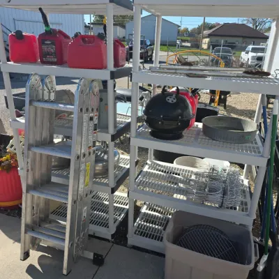 Sale Photo Thumbnail #140: Pair of white plastic shelving units holding gas cans, a portable kettle grill, metal pans, and lawn tools alongside an aluminum folding ladder.