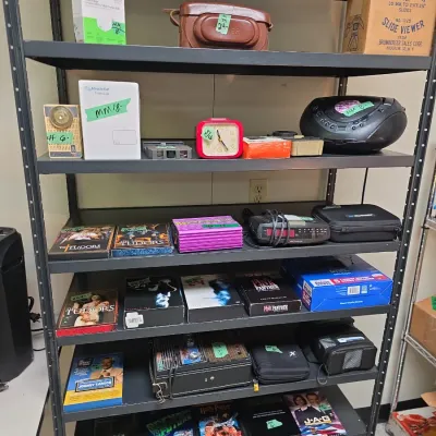 Sale Photo Thumbnail #47: Black metal shelving unit filled with assorted electronics, DVD box sets, video games, a retro Brumberger slide viewer, and a portable boombox.