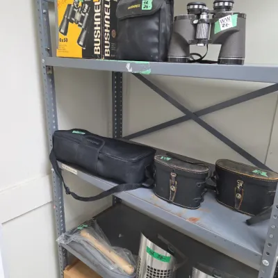 Sale Photo Thumbnail #74: A metal utility shelf holding various outdoor gear, including Bushnell 10x50 binoculars with original box and soft case, several hard cases, a small hammer, and metal sand scoops.