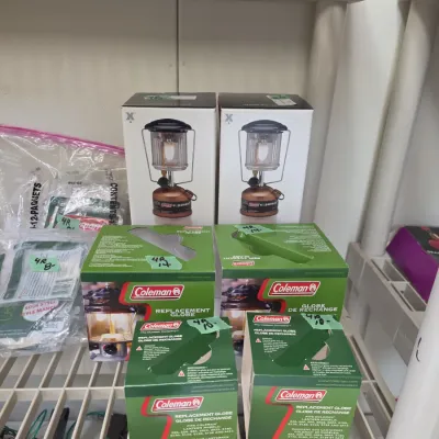 Sale Photo Thumbnail #52: Selection of Coleman camping supplies including replacement lantern globes and Exponent lanterns in original boxes, displayed on wire shelving.
