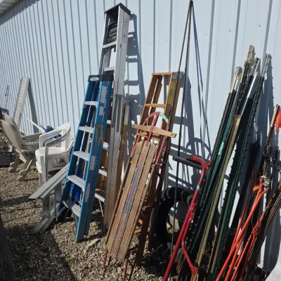Sale Photo Thumbnail #141: Assortment of outdoor equipment including a vintage wooden Flexible Flyer sled, fiberglass and aluminum step ladders, metal T-posts, and lawn chairs leaning against a corrugated metal wall.