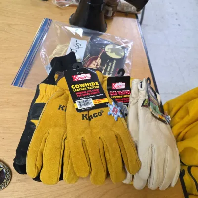 Sale Photo Thumbnail #62: Assortment of work gloves, including Kinco cowhide leather thermal driver gloves with tags, alongside a black Realistic portable megaphone on a wooden table.