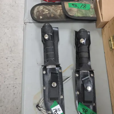 Sale Photo Thumbnail #79: Pair of military-style fixed-blade knives in black leather sheaths alongside boxed multi-tools and outdoor accessories.