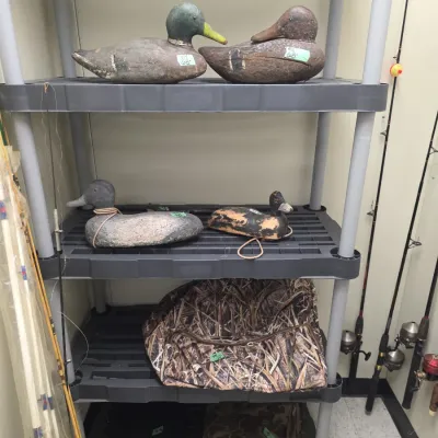 Sale Photo Thumbnail #67: Collection of vintage carved wooden duck decoys and hunting gear arranged on a plastic shelving unit alongside fishing rods.