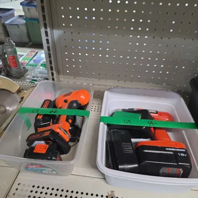 Sale Photo Thumbnail #137: Two plastic bins containing Black & Decker cordless power drills with rechargeable battery packs and chargers, sitting on store shelving.