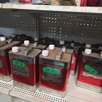 Sale Photo Thumbnail #134: Multiple vintage red Coleman 1-gallon camping fuel metal cans stored on store shelving.