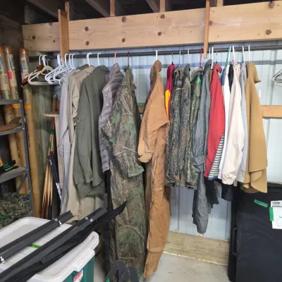Sale Photo Thumbnail #131: Collection of men's outerwear hanging on a rack, including camouflage hunting gear, coveralls, trench coats, and jackets.