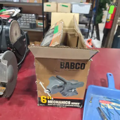 Sale Photo Thumbnail #110: Vintage Babco 6-inch Mechanics Series bench vise in its original cardboard box, set on a workshop table next to a bench grinder.