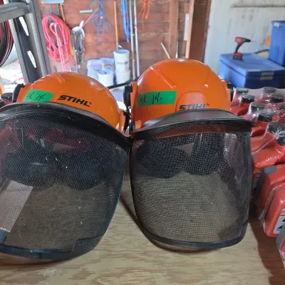 Sale Photo Thumbnail #127: Pair of orange STIHL safety hard hats featuring attached wire mesh face shields for outdoor power equipment work.