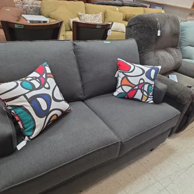 Sale Photo Thumbnail #5: Contemporary dark gray upholstered loveseat accented with two colorful abstract-pattern throw pillows, displayed alongside other living room seating in a showroom setting.