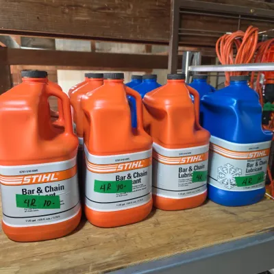 Sale Photo Thumbnail #125: Multiple one-gallon jugs of Stihl bar and chain lubricant, including standard orange and winter grade blue formulas, arranged on a garage shelf.