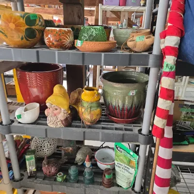 Sale Photo Thumbnail #115: Selection of garden décor and planters on a black plastic shelving unit, including ceramic pots, gnome figurines, bird statues, glass insulators, and lawn fertilizer.
