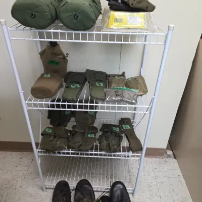 Sale Photo Thumbnail #86: Collection of military surplus gear displayed on a white wire shelving unit, including canteens with covers, ammunition pouches, webbing, and leather combat boots.