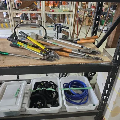 Sale Photo Thumbnail #113: Assortment of long-handled loppers and garden shears displayed on a shelving unit above plastic tubs filled with hoses.