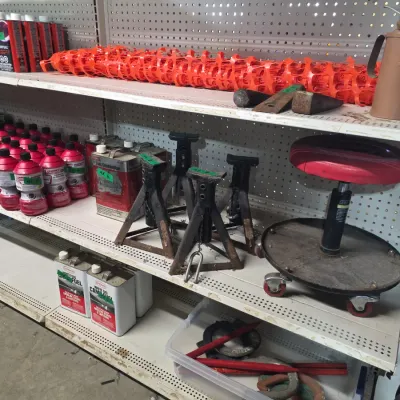 Sale Photo Thumbnail #132: Retail shelving containing workshop tools and equipment, including jack stands, a rolling mechanic's stool, fuel cans, and safety netting.