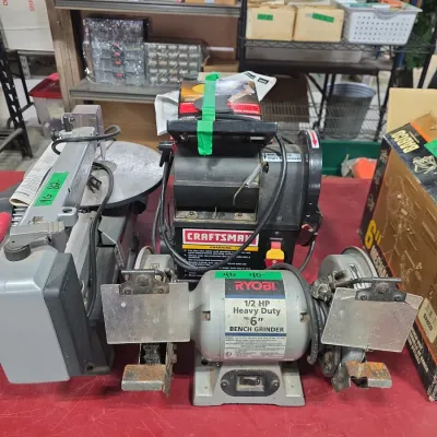 Sale Photo Thumbnail #109: Assorted power tools including a Ryobi 1/2 HP 6-inch bench grinder, Craftsman belt sander, and a scroll saw displayed on a workbench.