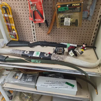 Sale Photo Thumbnail #66: Collection of sporting goods including mounted longhorn steer horns, a green recurve bow, an air rifle, fishing reels, and vintage packaged tools on pegboard shelves.