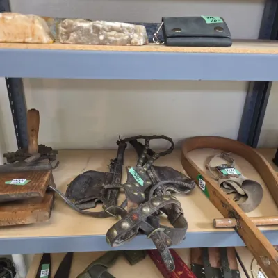 Sale Photo Thumbnail #60: Collection of vintage tools and collectibles including brass-studded leather horse tack, a wooden hand-crank machine, shear blades, and a small hatchet displayed on metal shelving.