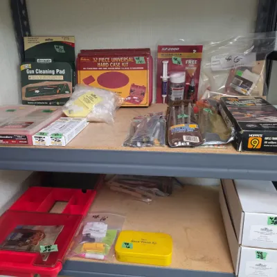 Sale Photo Thumbnail #85: Collection of gun maintenance and cleaning supplies, including universal kits, cleaning pads, solvents, and cleaning rods displayed on utility shelving.